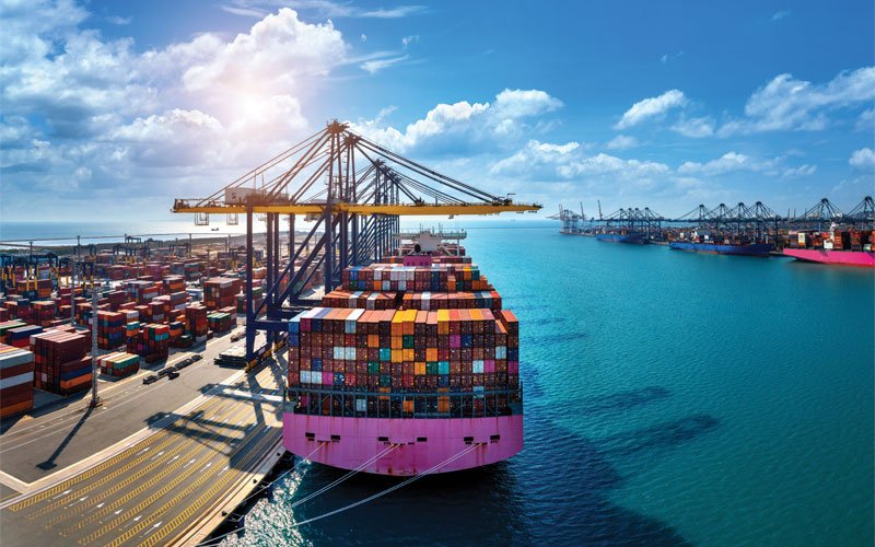 an aerial view of a busy cargo port. A large cargo ship is docked at the port, with its deck stacked high with multicolored shipping containers.